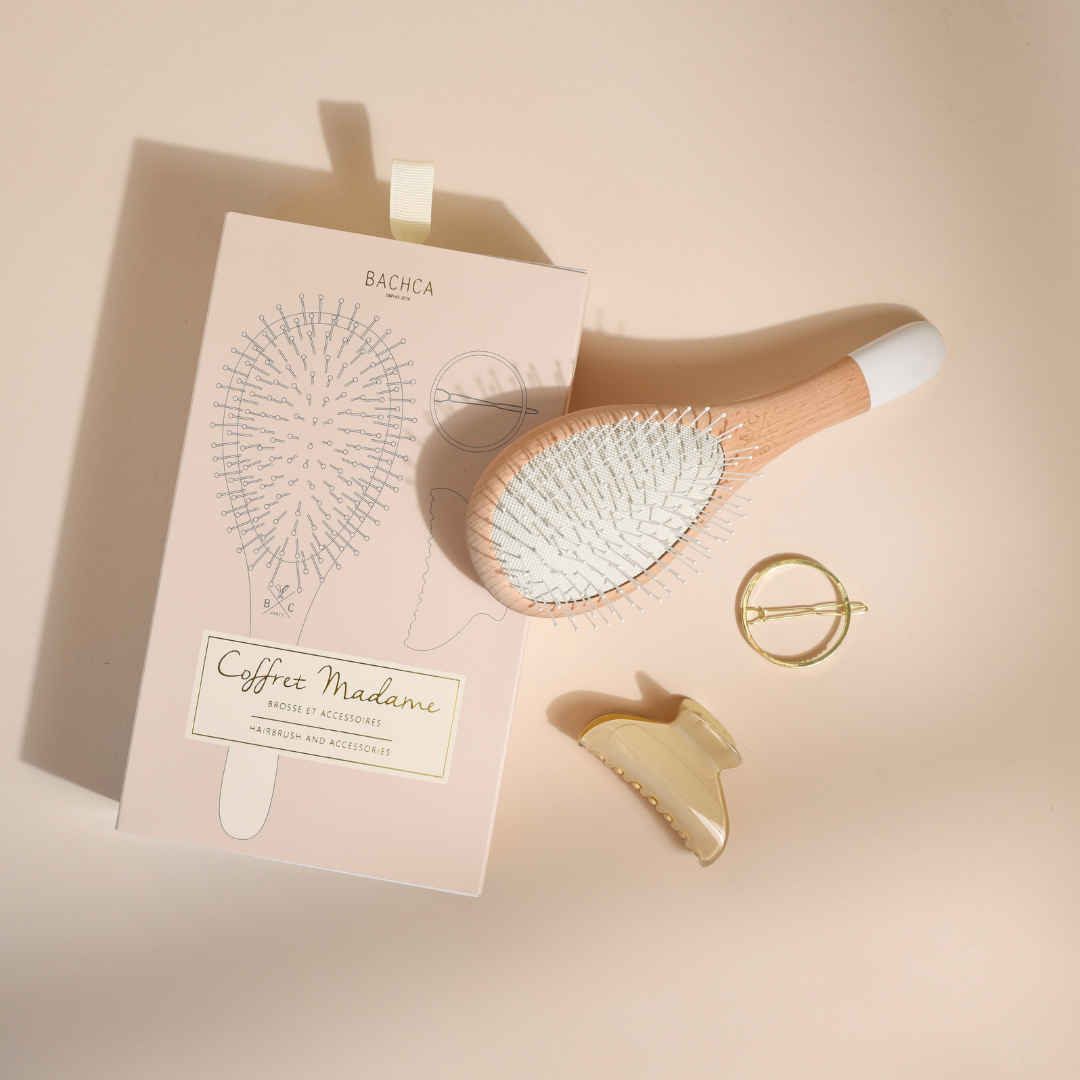 Coffret Madame Bachca avec brosse en bois à picots nylon, pince crème et barrette ronde dorée, présenté à plat sur fond beige à côté de son étui illustré.