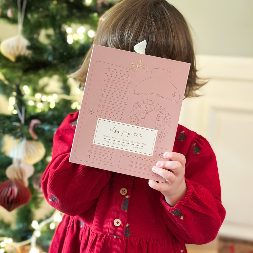 Petite fille qui tient le coffret les pépites bois de rose de la marque Bachca et cache son visage, en arrière plan on peut voir un sapin de noël.