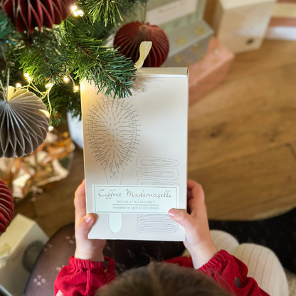 Une petite fille tient le coffret Mademoiselle de la marque Bachca dans ses mains sous un sapin de noël.