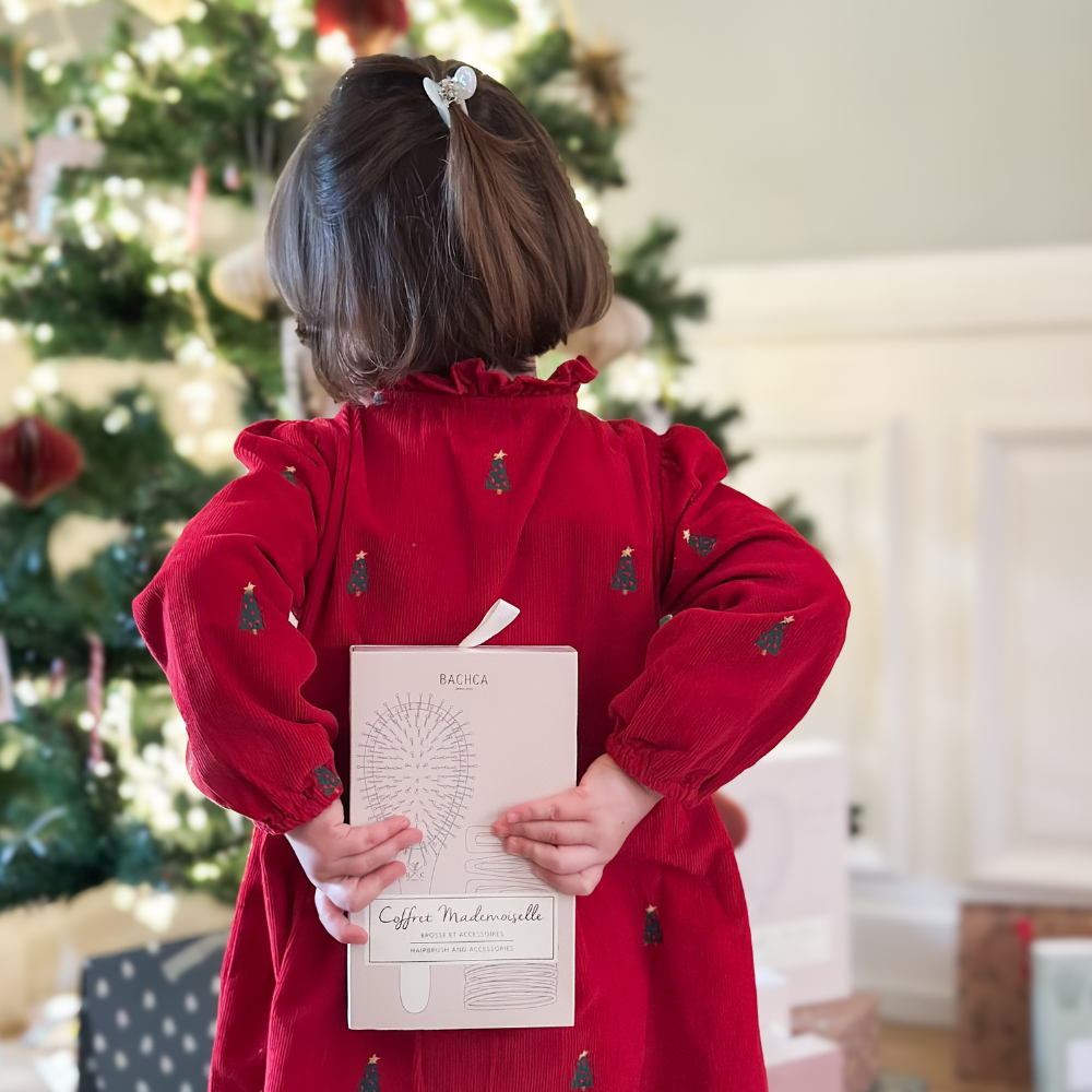 Petite fille de dos face à un sapin qui tient un coffret de la marque Bachca dans son dos