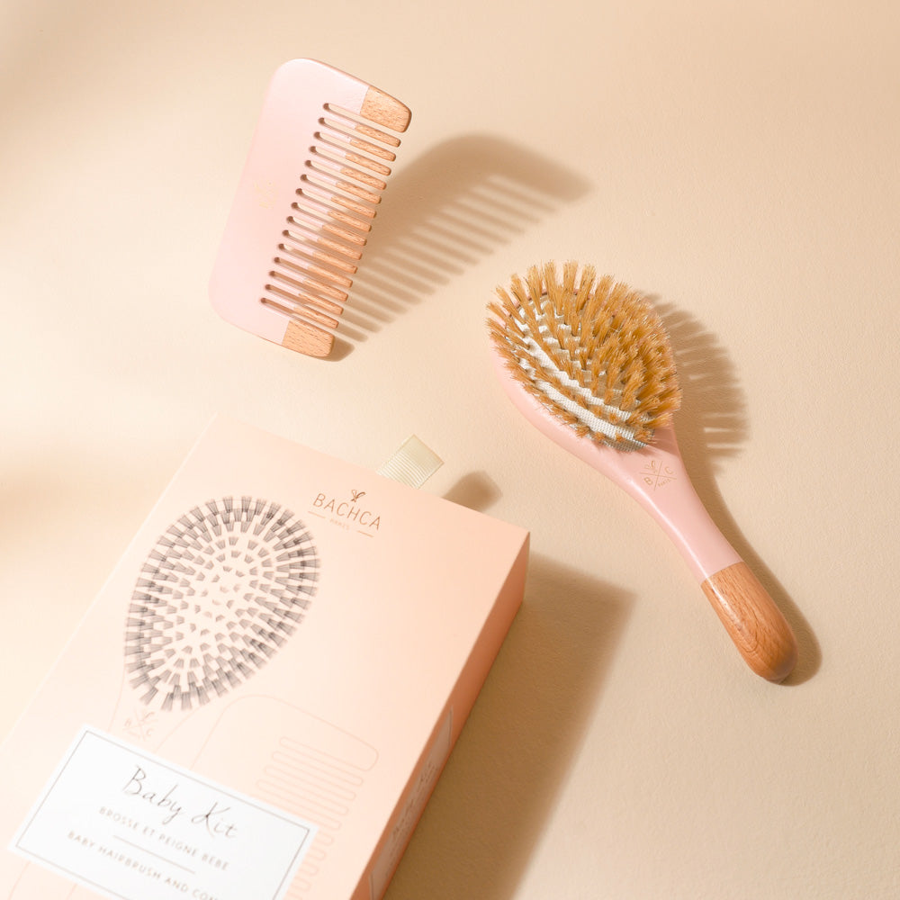 Kit naissance Bachca présenté sur un fond beige. À côté de la boîte rose poudré, on aperçoit une petite brosse en bois et poils doux au manche rose ainsi qu’un peigne assorti, aux détails en bois naturel. La lumière met en relief les ombres délicates des accessoires.