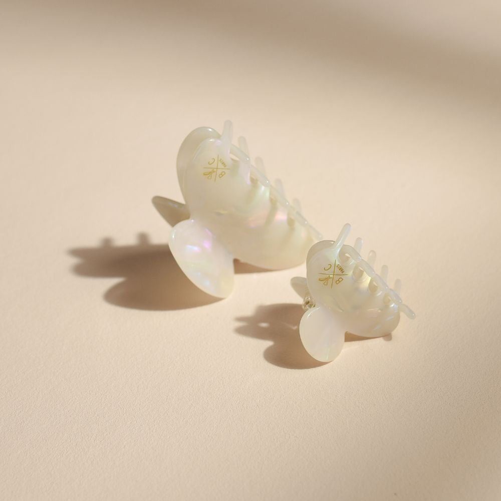 Deux pinces à cheveux Bachca en acétate nacré, posées sur fond beige, aux reflets irisés et finitions dorées.