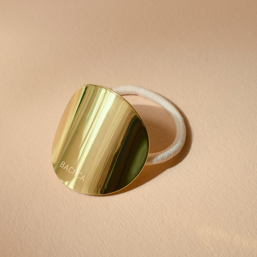 Élastique à cheveux blanc avec une plaque ronde dorée de la marque Bachca. Le produit est sur fond rose pâle.