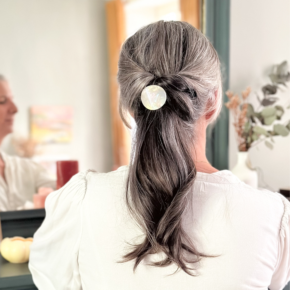 Femme de dos aux cheveux gris. Elle porte dans ses cheveux un élastique plaque nacre, on voit son reflet dans un miroir.