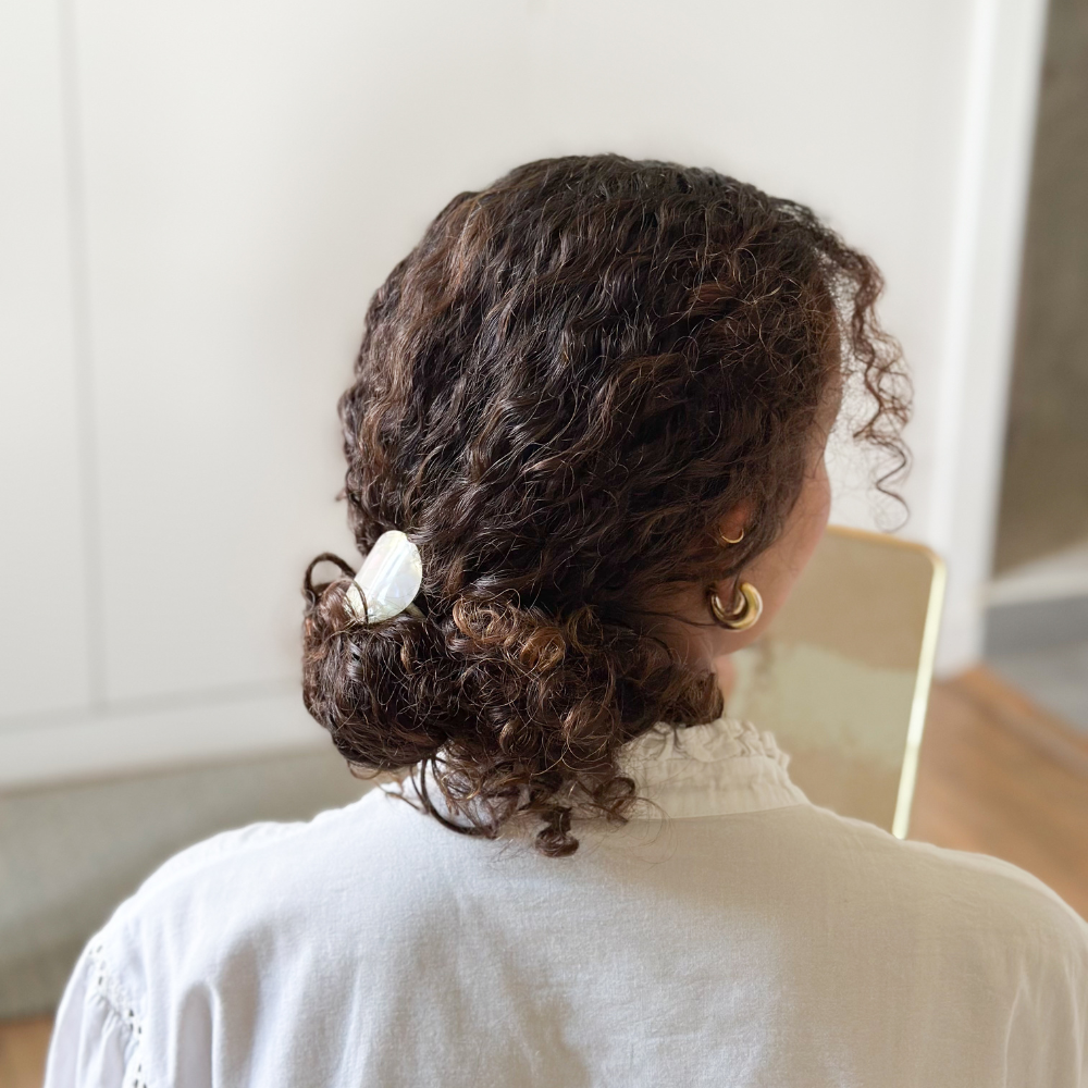 Femme de dos aux cheveux bouclés. Elle porte dans ses cheveux un élastique plaque nacre en chignon bas.