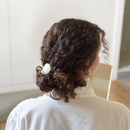 Femme de dos aux cheveux bouclés. Elle porte dans ses cheveux un élastique plaque nacre en chignon bas.