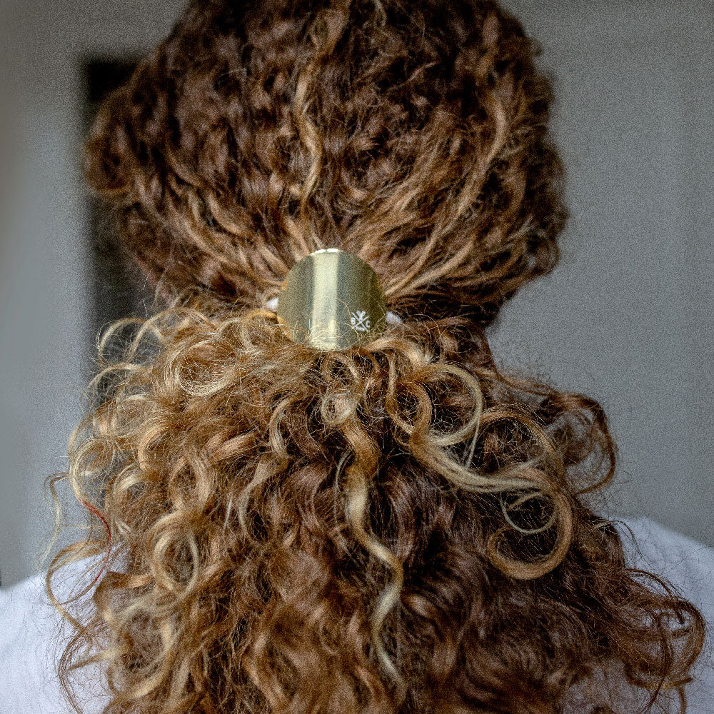 Cheveux bouclés attachés avec un élastique plaque or métallique Bachca, formant une demi-queue élégante vue de dos.