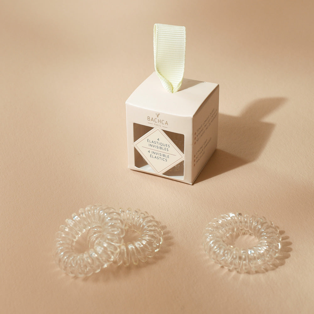 Boîte d’élastiques invisibles Bachca avec trois élastiques transparents en spirale placés devant, photographiée sur une surface beige.