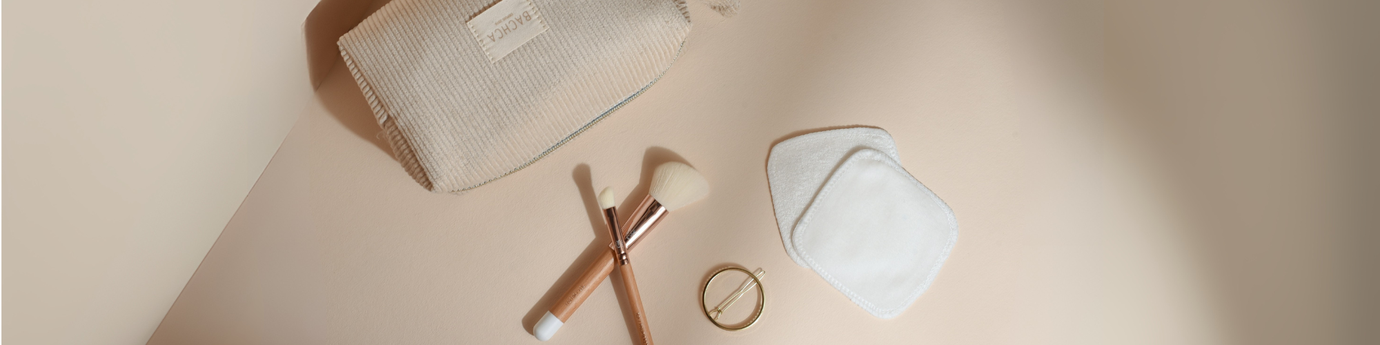 Trousse makeup Bachca avec cotons démaquillants réutilisables, deux pinceaux maquillage teint et yeux en bois, barrette ronde dorée. Produits présentés sur fond rose pâle.