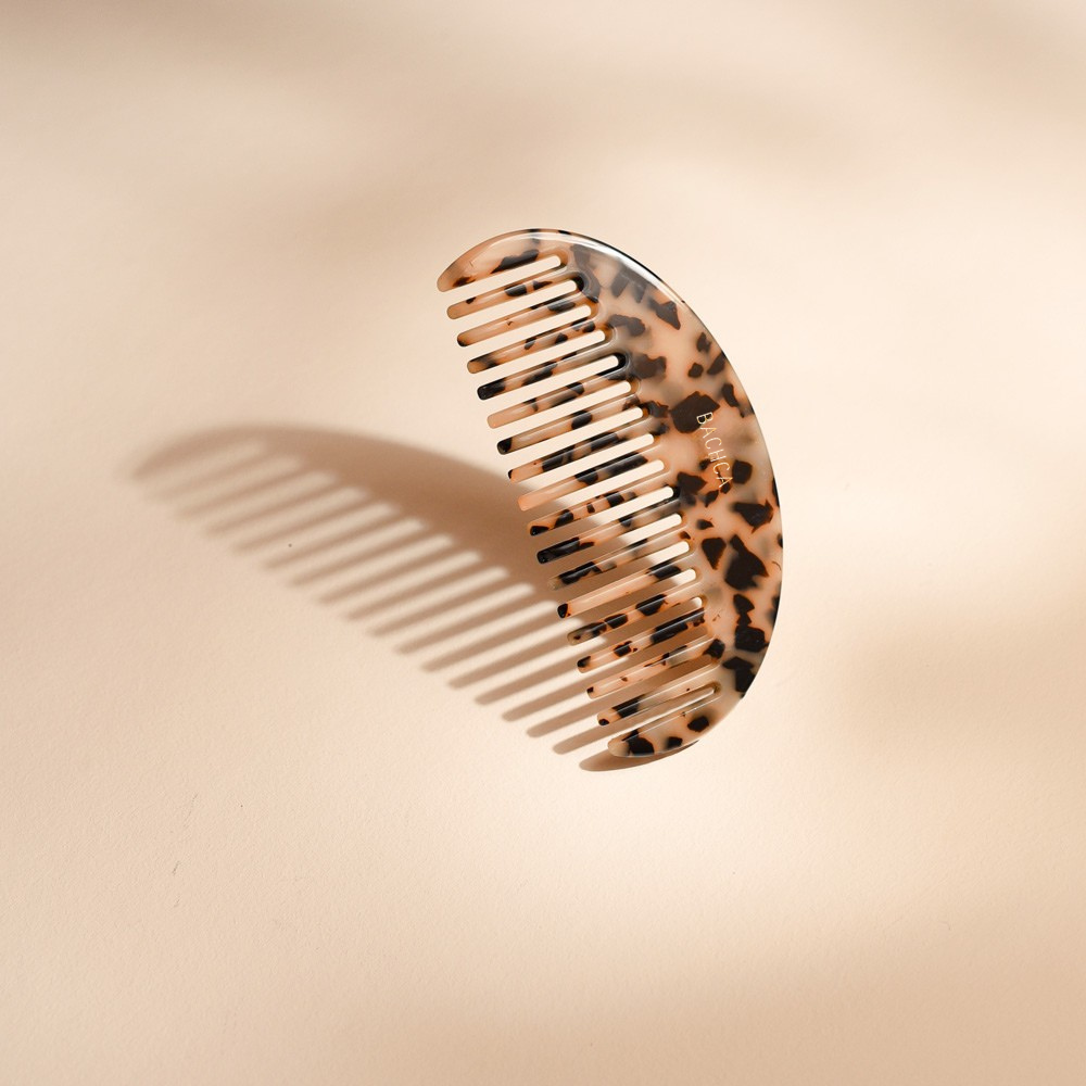 Peigne à cheveux en acétate motif écaille, dents larges et forme légèrement arrondie, signé Bachca, présenté sur fond rose pâle.