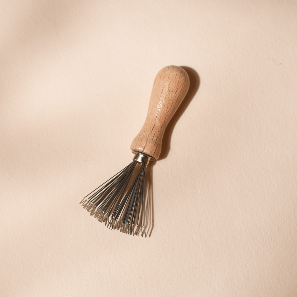 Peigne nettoyant Bachca en métal et bois brut, idéal pour entretenir les brosses à cheveux. Produit sur fond rose pâle.