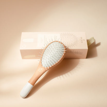 Brosse à cheveux Bachca petit format pour apporter volume et démêlage, en bois de hêtre naturel avec picots souples, posée sur sa boîte beige, sur fond sable.