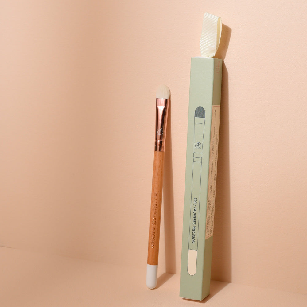 Pinceau à paupières précision Bachca avec manche en bois et poils doux, présenté debout à côté de son emballage vert pastel, photographié sur une surface beige.