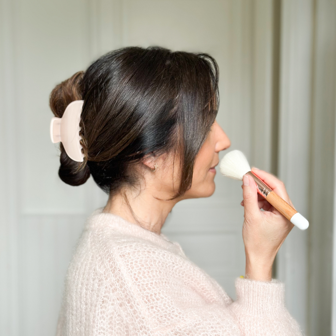 Femme appliquant de la poudre avec un pinceau teint Bachca en bois, cheveux relevés avec une grande pince à cheveux rose, en intérieur lumineux.