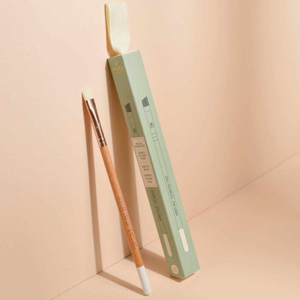 Pinceau sourcils et eye-liner Bachca avec manche en bois et embout biseauté, présenté debout contre son étui vert pastel, photographié sur une surface beige.