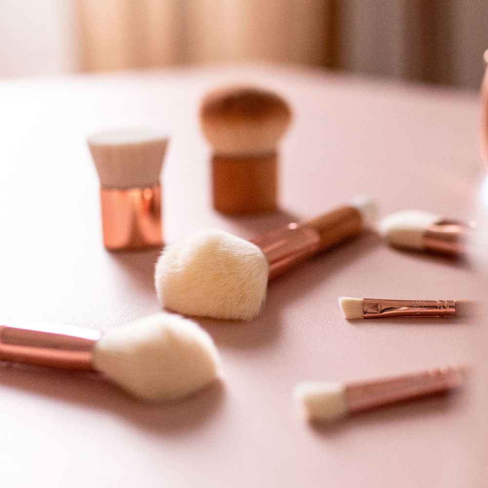Set de pinceaux de maquillage Bachca aux manches bois et embouts rose cuivré, disposés sur une surface rose poudré.