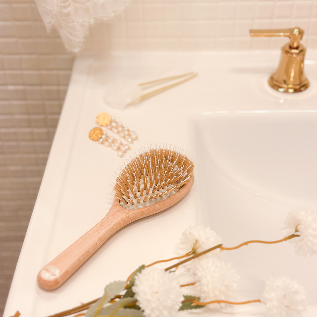 Brosse à cheveux Bachca en bois posée au bord d’un lavabo, entourée de fleurs blanches, bijoux et accessoires dorés.