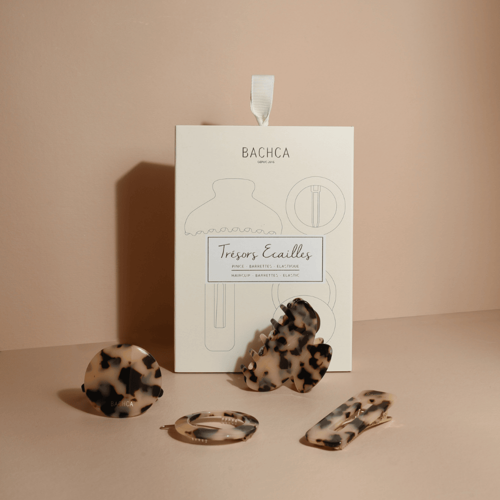 Coffret d’accessoires cheveux en écaille de la marque Bachca : pince, barrette, barrette ronde et élastique plaque, présentés sur fond rose pâle.
