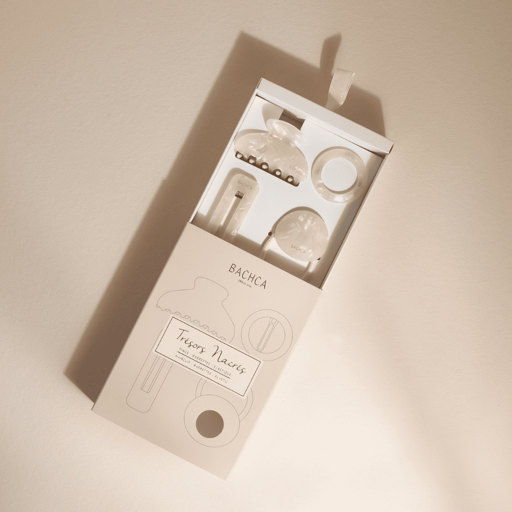 Coffret d’accessoires cheveux en nacre de la marque Bachca : pince, barrette, barrette ronde et élastique plaque, présentés sur fond rose pâle.