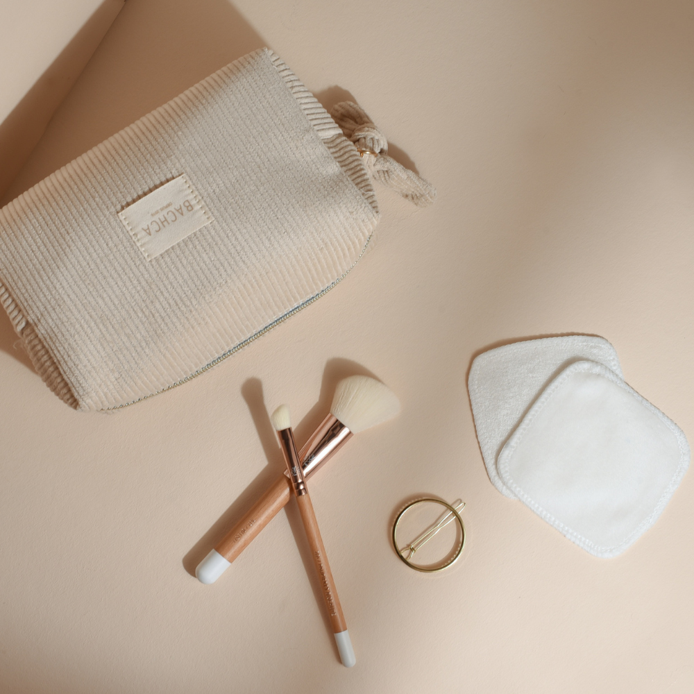 Trousse makeup Bachca avec cotons démaquillants réutilisables, pinceaux en bois, barrette dorée ronde et pinceaux yeux et teint à poils blancs. Produits présentés sur fond rose pâle.