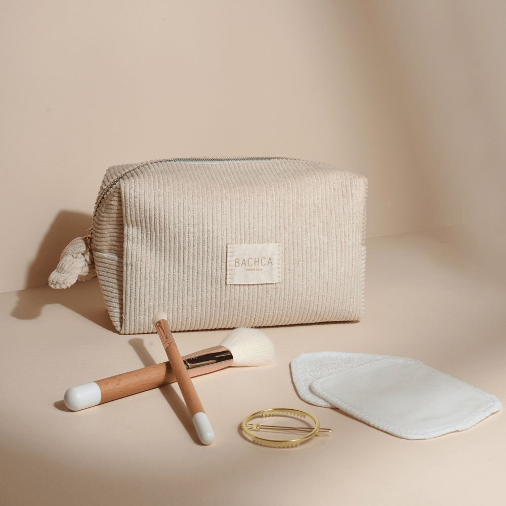 Trousse makeup en velours côtelé beige Bachca avec cotons démaquillants réutilisables, pinceaux en bois, barrette dorée ronde et pinceaux yeux et teint à poils blancs. Produits présentés sur fond rose pâle.