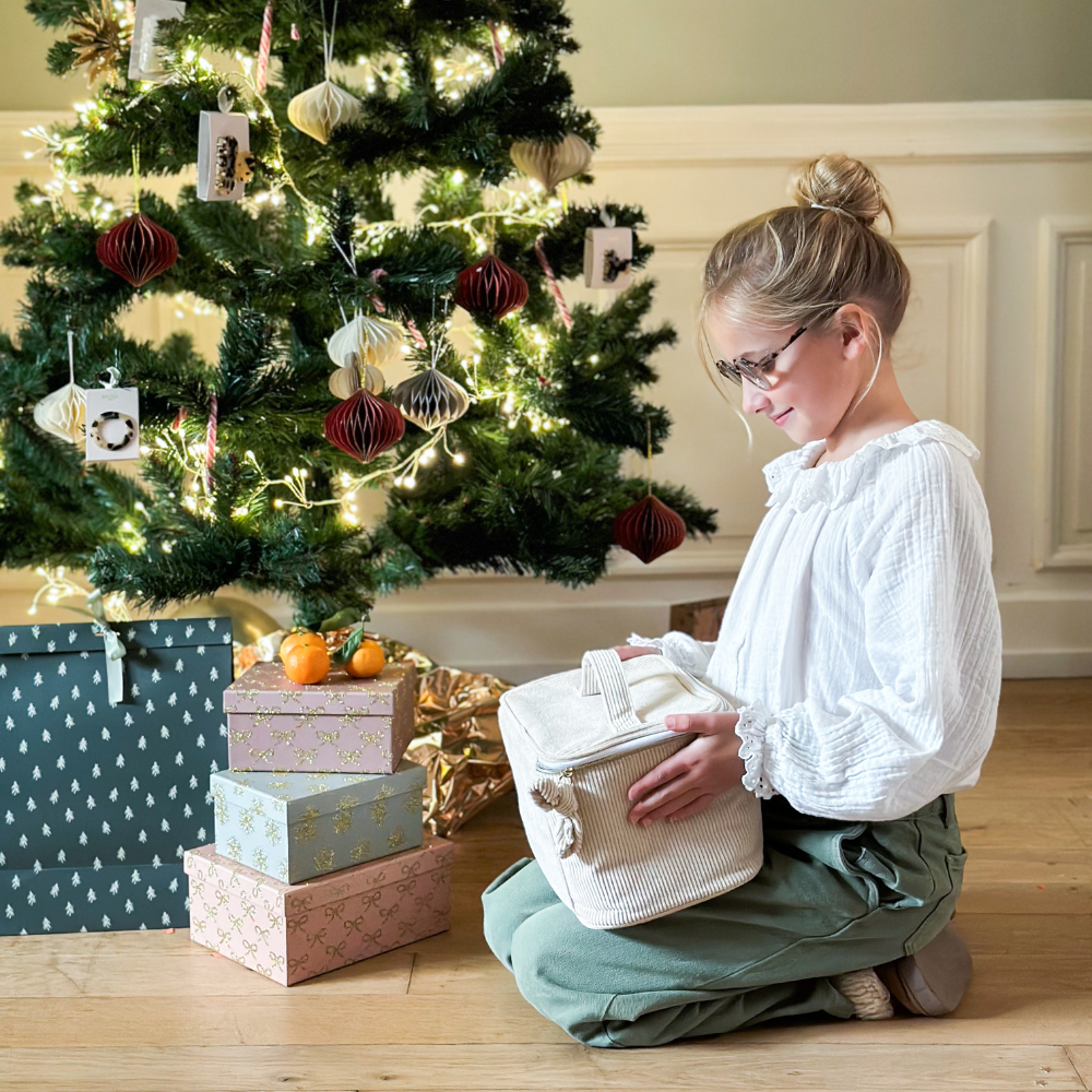 Une jeune fille est assise près d'un sapin de noël, elle tient dans ses mains un vanity en velours côtelé beige de la marque Bachca.