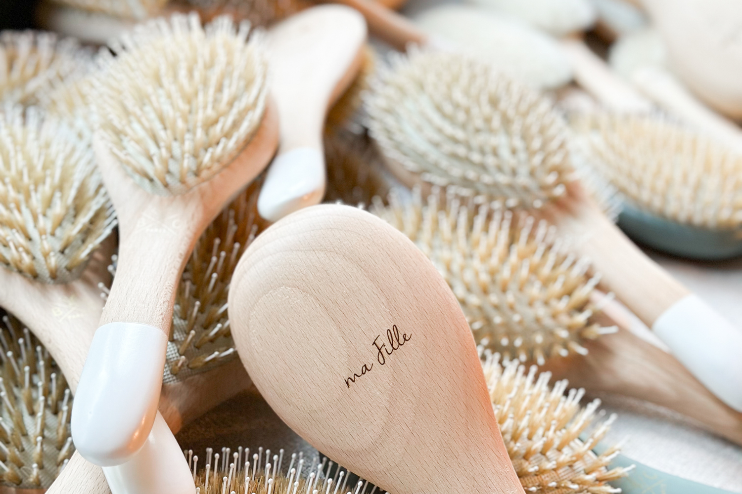 Gros plan sur une brosse à cheveux gravée "ma fille" de la marque Bachca, plusieurs brosses en vrac en fond.