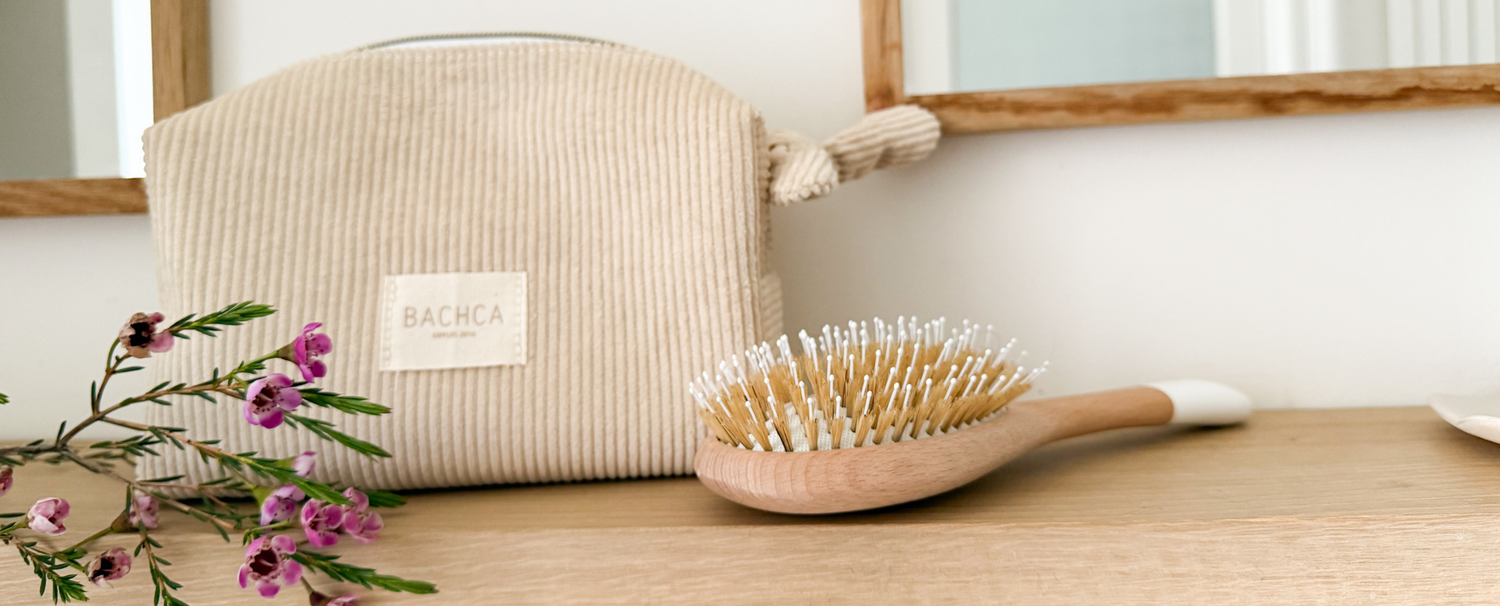Une trousse maquillage en velours côtelé beige et une brosse à cheveux de la marque Bachca sont disposées sur une étagère de salle de bain.