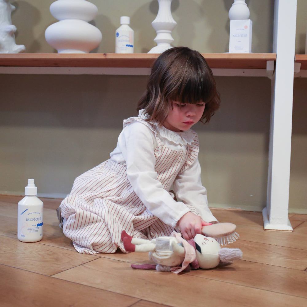 Petite fille assise, elle joue avec son doudou et le brosse avec soin grâce à une brosse à cheveux de la marque Bachca.
