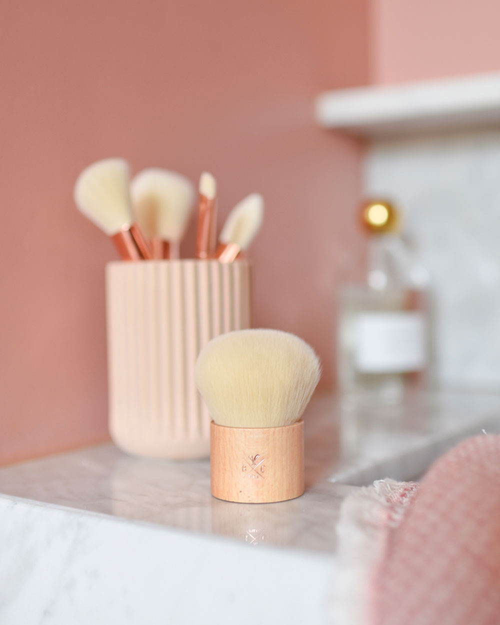 Pinceau kabuki Bachca à manche bois clair posé sur un plan de salle de bain en marbre, avec un pot de pinceaux de maquillage en arrière-plan.