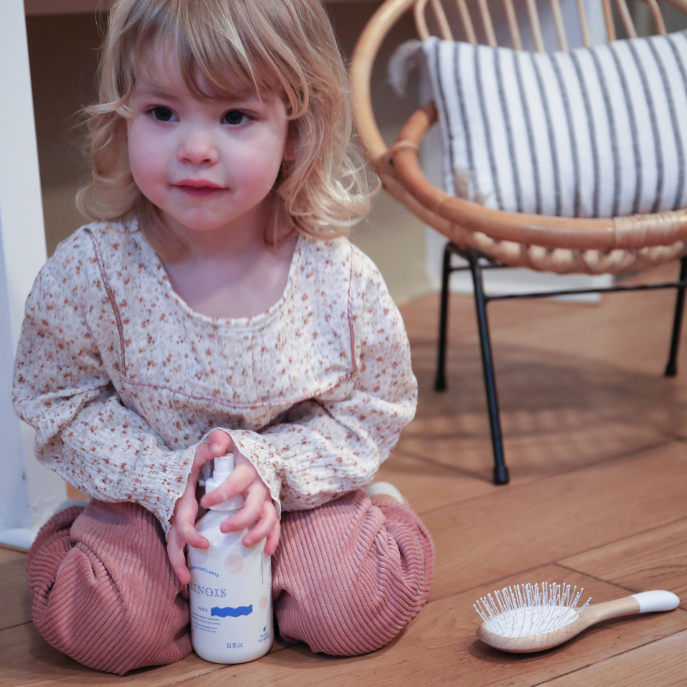 Petite fille assise qui our avec une brûme pour les cheveux dans ses mains, on peut observer une brosse à cheveux de la marque Bachca posée à côté d&