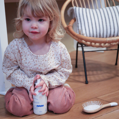 Petite fille assise qui our avec une brûme pour les cheveux dans ses mains, on peut observer une brosse à cheveux de la marque Bachca posée à côté d&