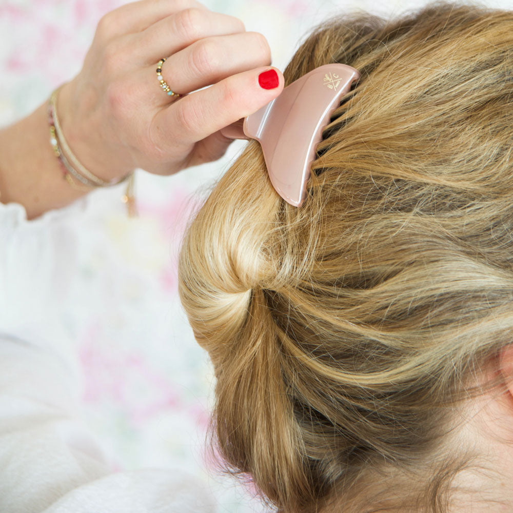 Femme attachant ses cheveux blonds avec une pince à cheveux Bachca terracotta en acétate de cellulose, accessoire élégant et pratique pour chignon ou demi-queue.