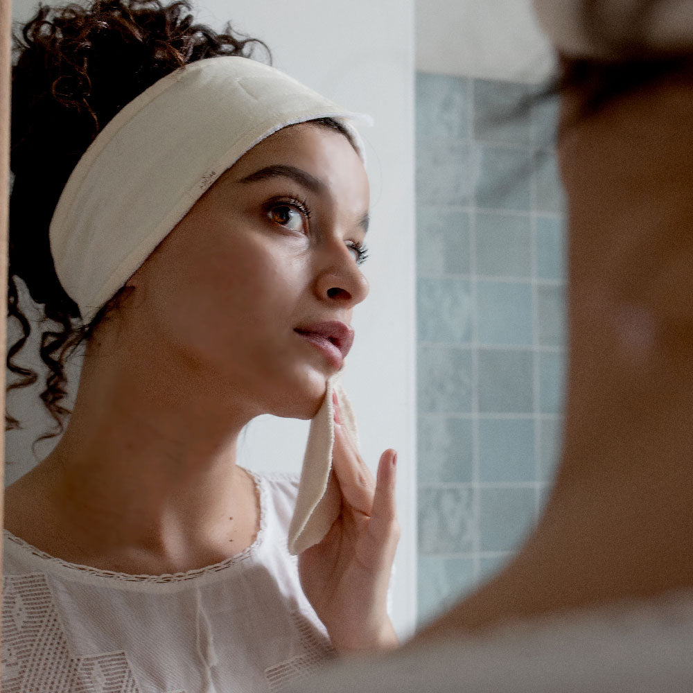 Femme portant un bandeau écru Bachca pour le nettoyage du visage, utilisant un coton démaquillant réutilisable devant un miroir dans une salle de bain.