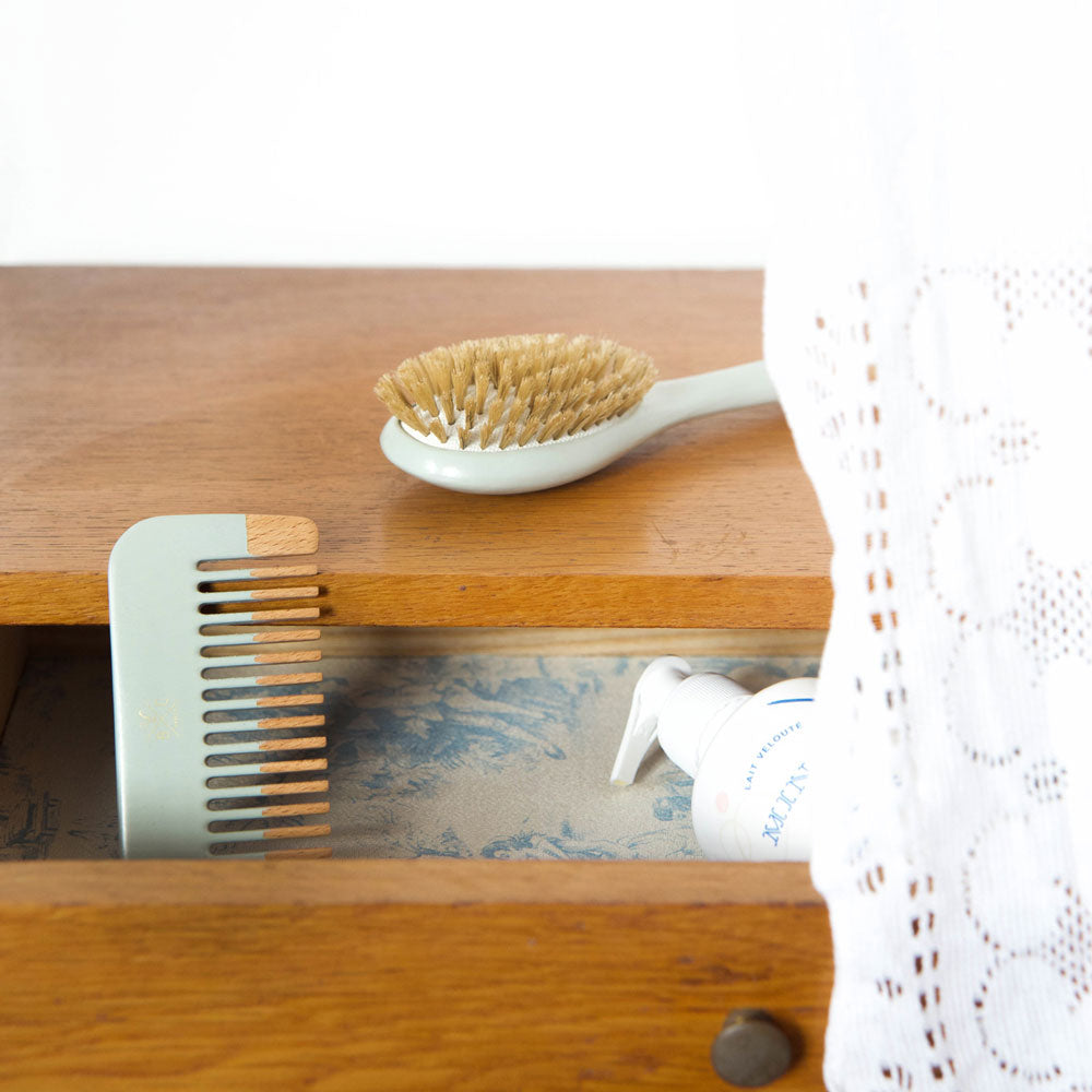 Une brosse bébé Bachca aux poils doux posée sur un meuble en bois et un petit peigne assorti en bois et teinte pastel rangé dans un tiroir entrouvert.