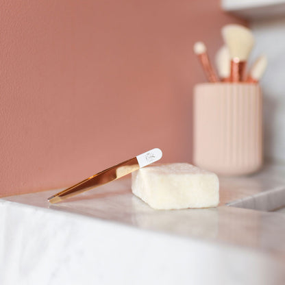 Pince à épiler dorée et blanche Bachca posée sur un lavabo en marbre, avec un savon solide à côté et un pot de pinceaux en arrière-plan.
