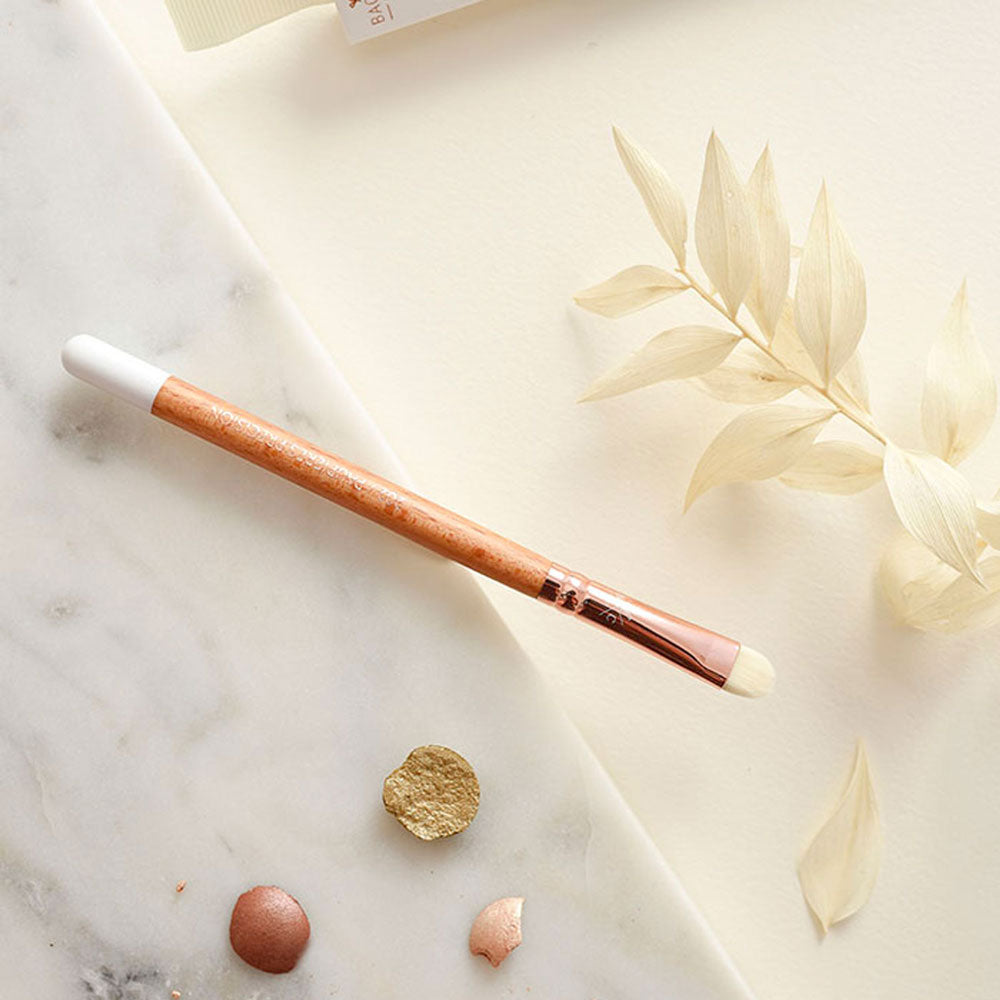 Pinceau précision pour paupières Bachca avec manche en bois et embout blanc, posé sur une surface en marbre avec des pigments de maquillage.