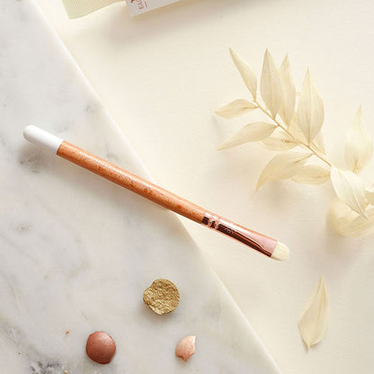 Pinceau précision pour paupières Bachca avec manche en bois et embout blanc, posé sur une surface en marbre avec des pigments de maquillage.